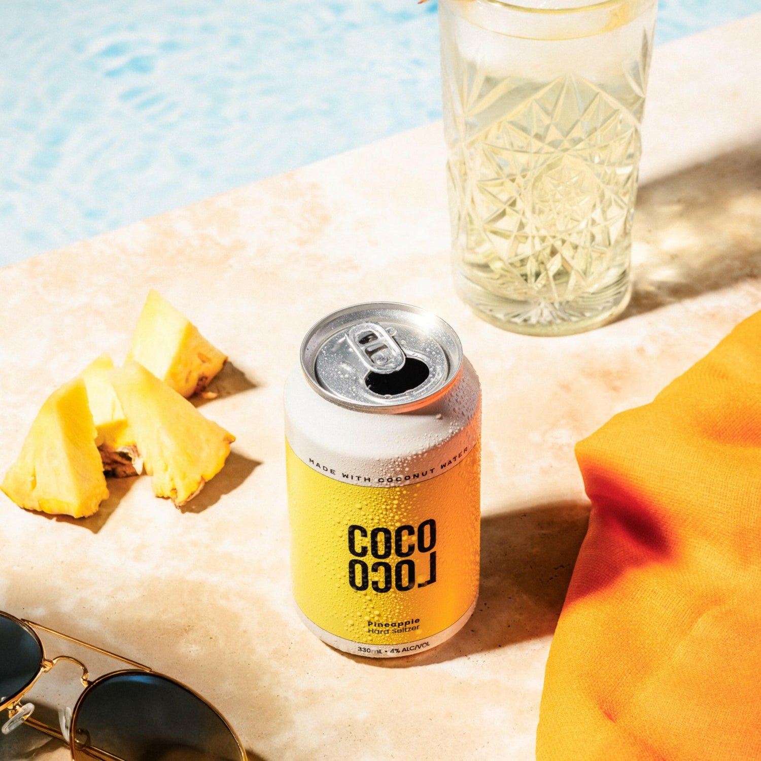 Coco Loco drink pineapple can with a elegant glass, pineapple slices, and sunglasses sat poolside.