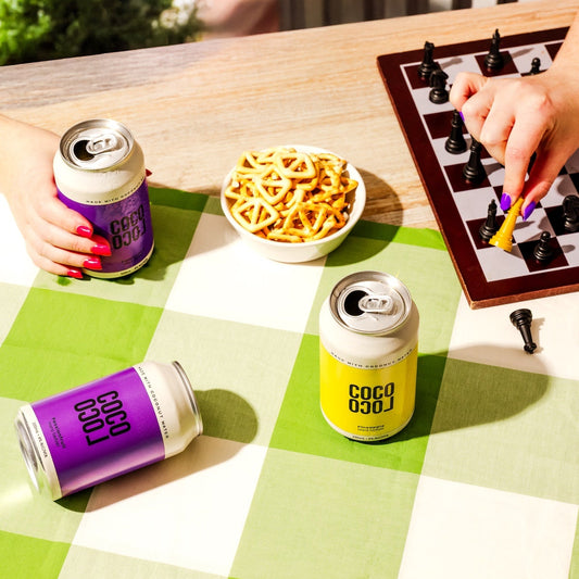 Two cans of Coco Loco drinks on a checkered tablecloth with snacks and a chessboard.