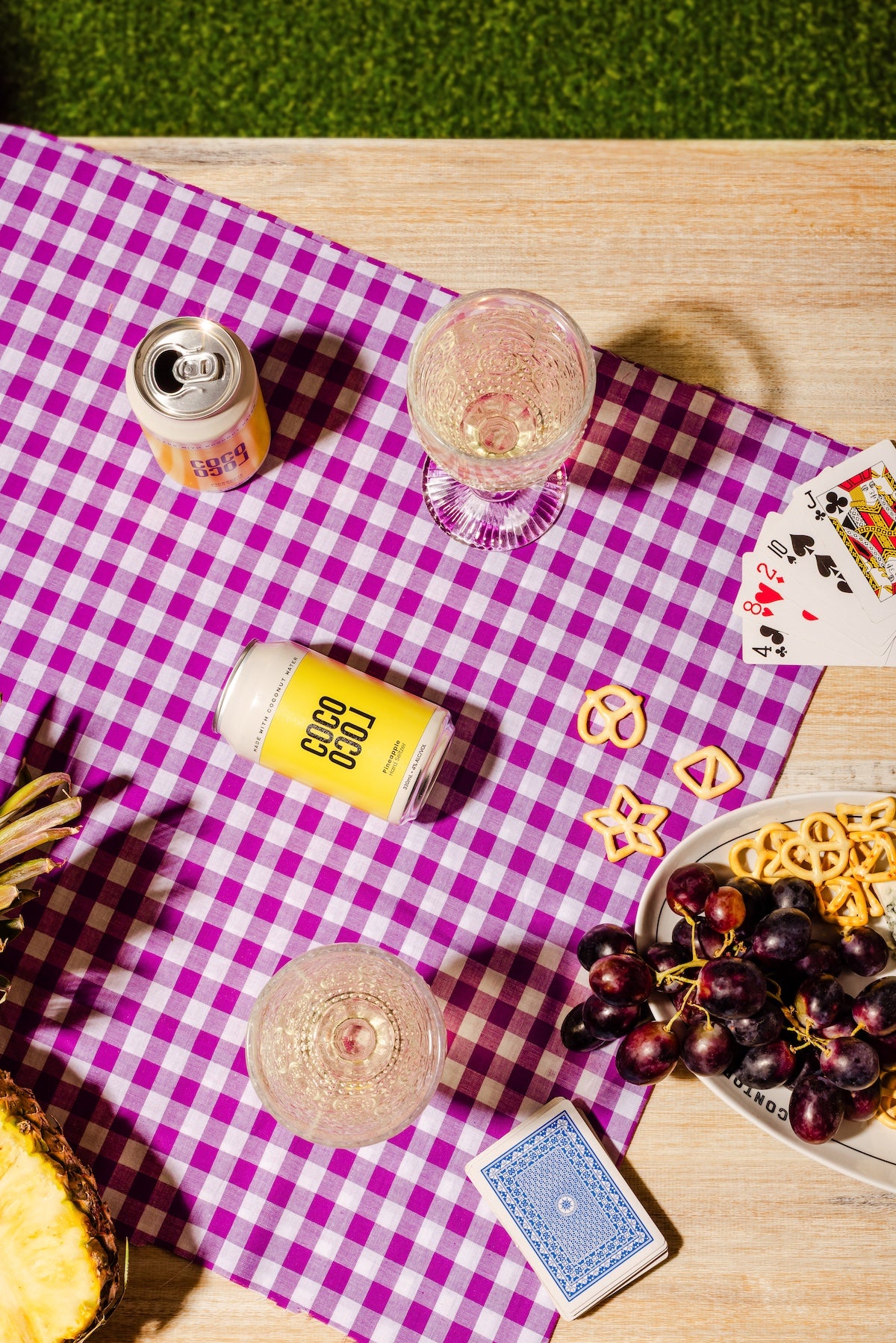 Picnic setup with Coco Loco drinks, snacks, and cards on a purple checkered tablecloth.