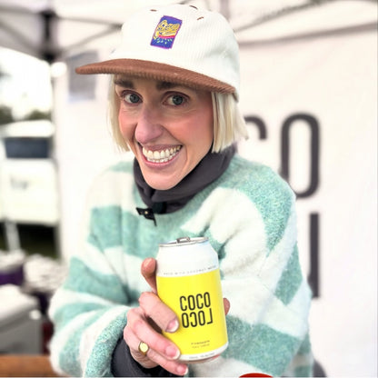 Coco Loco founder, Jill, wearing a Passiona corduroy cap and a mint-and-white striped sweater holds a yellow can of 'COCO LOCO'