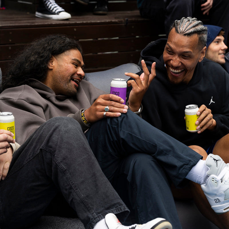 Two men sitting together, one holding a passionfruit hard seltzer can and the other a Coco Loco hard seltzer can.