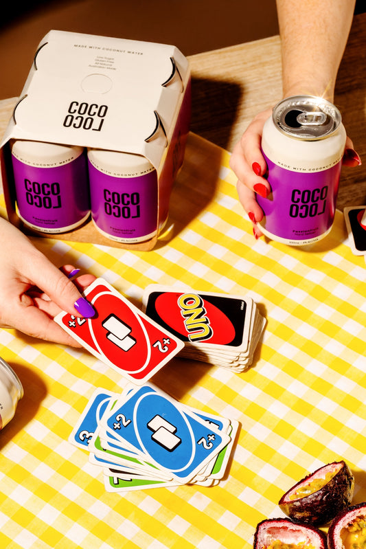 coco loco passionfruit seltzer cans on a yellow checkered tablecloth, surrounded by UNO playing cards and real passionfruit.