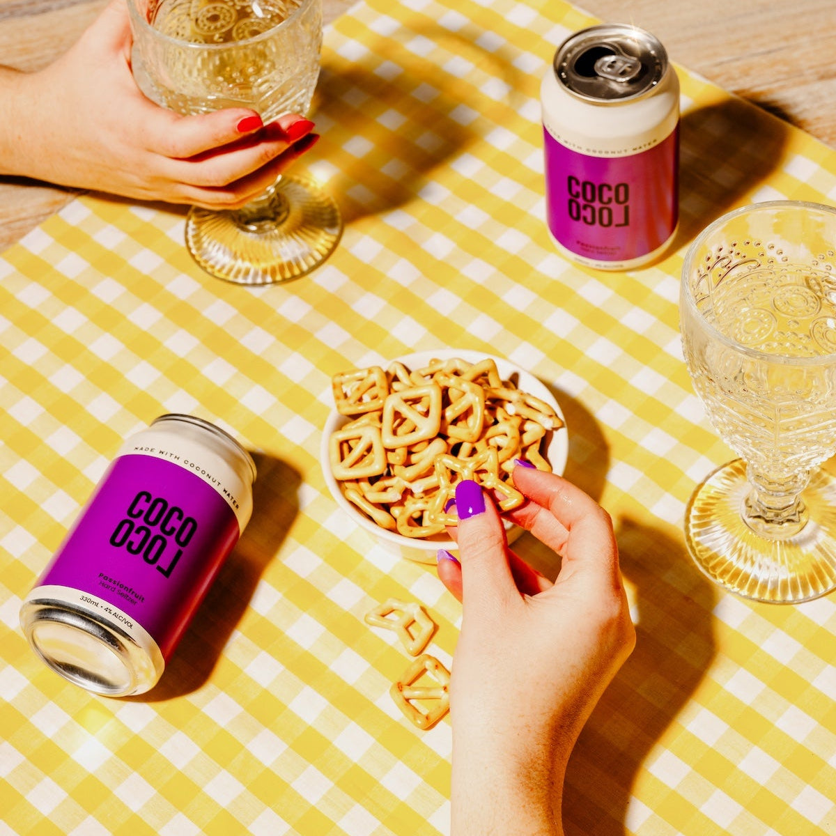 Two people enjoying drinks with Coco Loco alcoholic coconut water cans on a yellow checkered tablecloth.