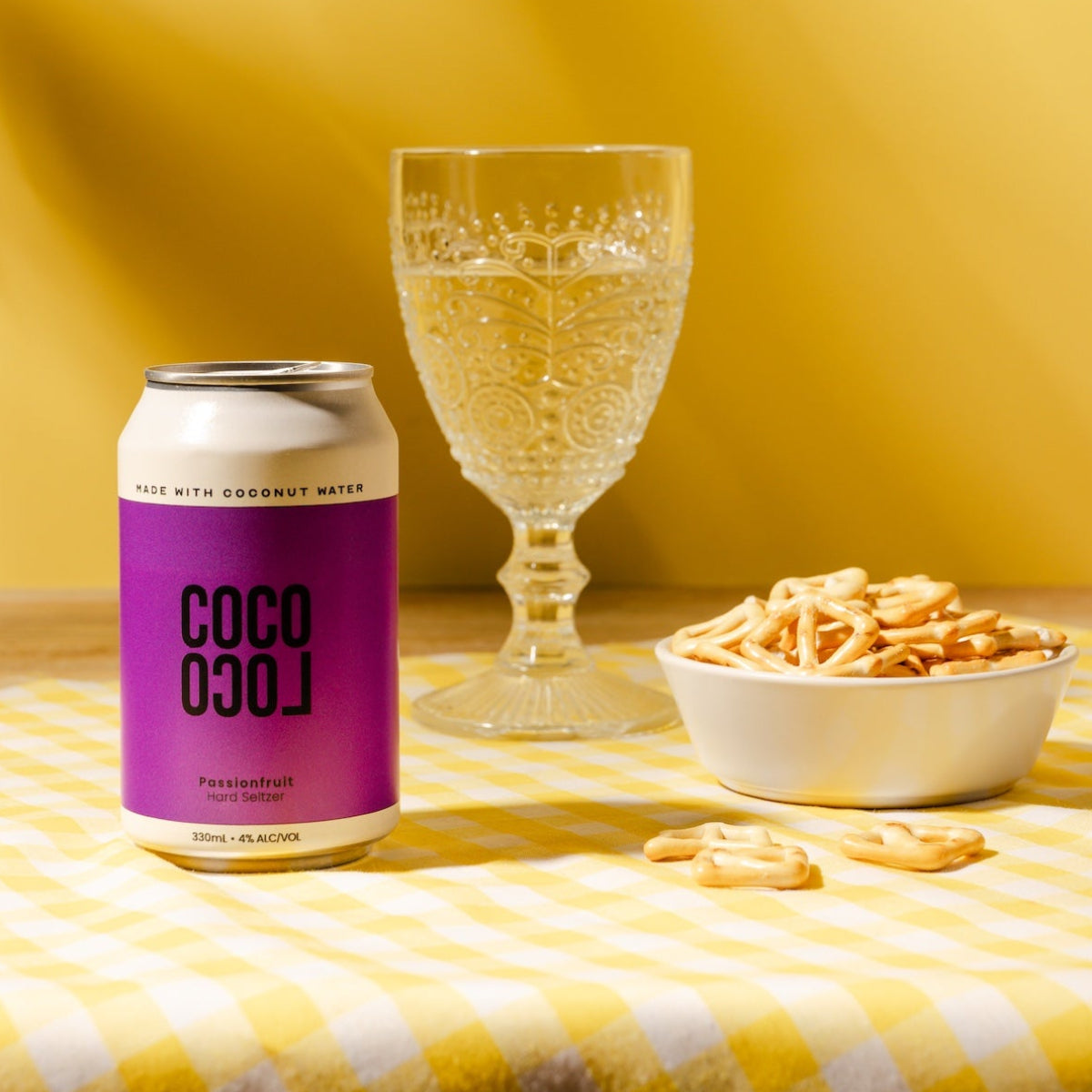 Coco Loco low sugar alcohol can with a glass of sparkling water and a bowl of snacks on a yellow checkered tablecloth.