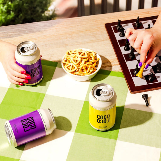 Two cans of coco drink on a checkered tablecloth with snacks and a chessboard.