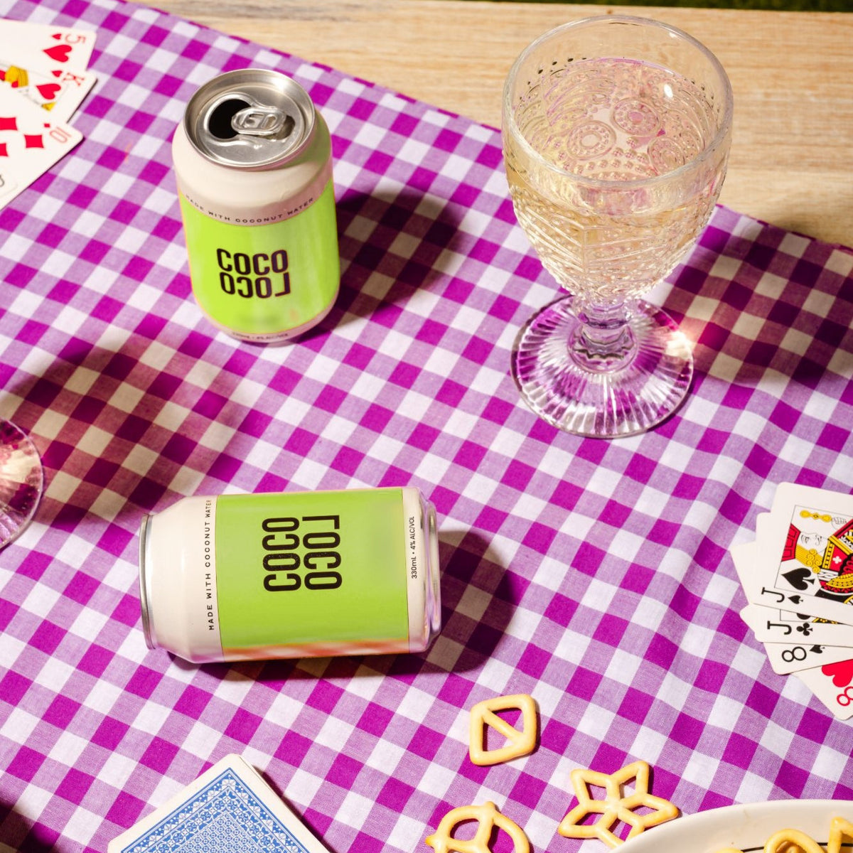 Two Coco Loco Margarita cans on a purple gingham picnic table with a glass, playing cards and pretzels