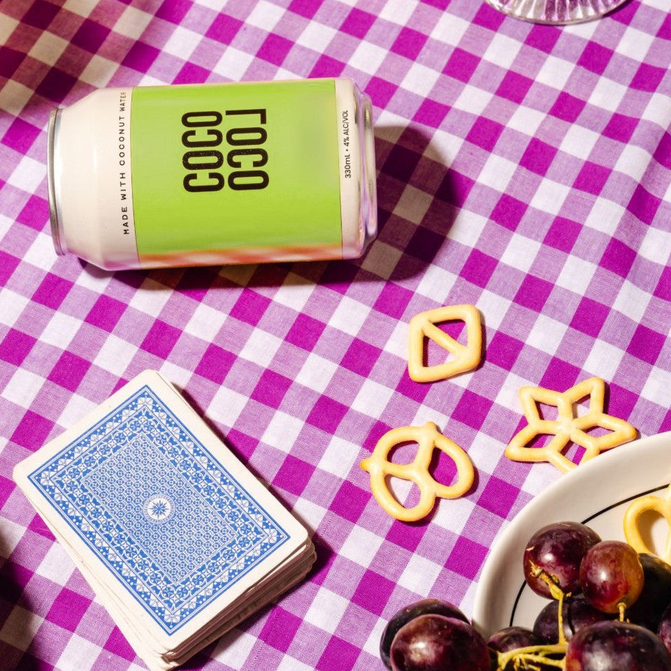 A Coco Loco Margarita can resting on a purple gingham tablecloth beside playing cards and pretzels